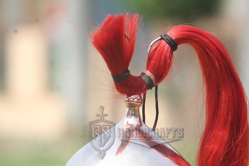 May include: Close-up of a silver and red helmet with a bright red plume. The plume is secured with black bands and a black cord. The helmet has a decorative crest and the text "RR HANDICRAFTS".