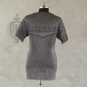 May include: A short-sleeved chainmail shirt displayed on a mannequin. The shirt is made of small, interlocking metal rings, creating a flexible, protective garment. The background is a light gray wall with a decorative emblem.