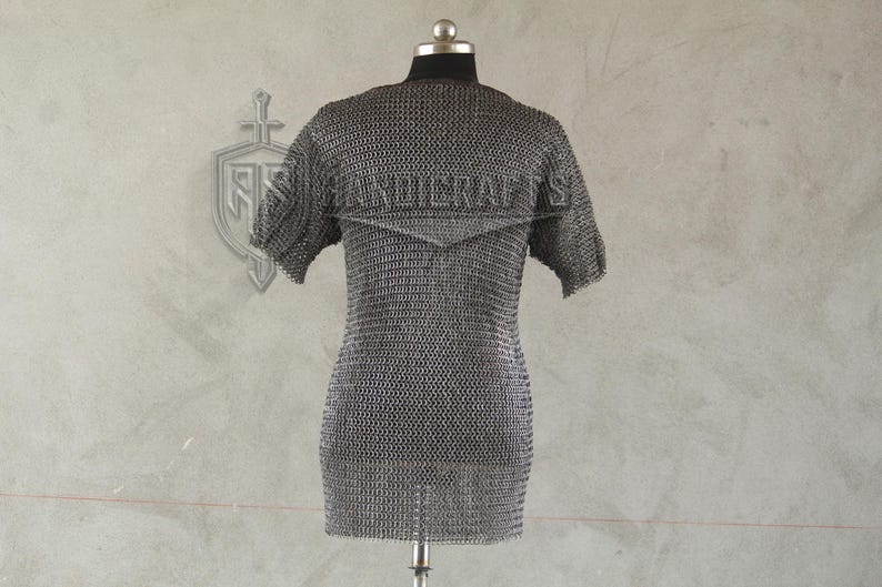 May include: A short-sleeved chainmail shirt displayed on a mannequin. The shirt is made of small, interlocking metal rings, creating a flexible, protective garment. The background is a light gray wall with a decorative emblem.