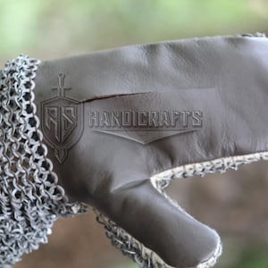 May include: Close-up of a medieval gauntlet. The gauntlet features a chainmail sleeve and a gray leather glove. The text "HANDICRAFTS" is visible on the leather. The gauntlet is designed for protection and historical reenactment.