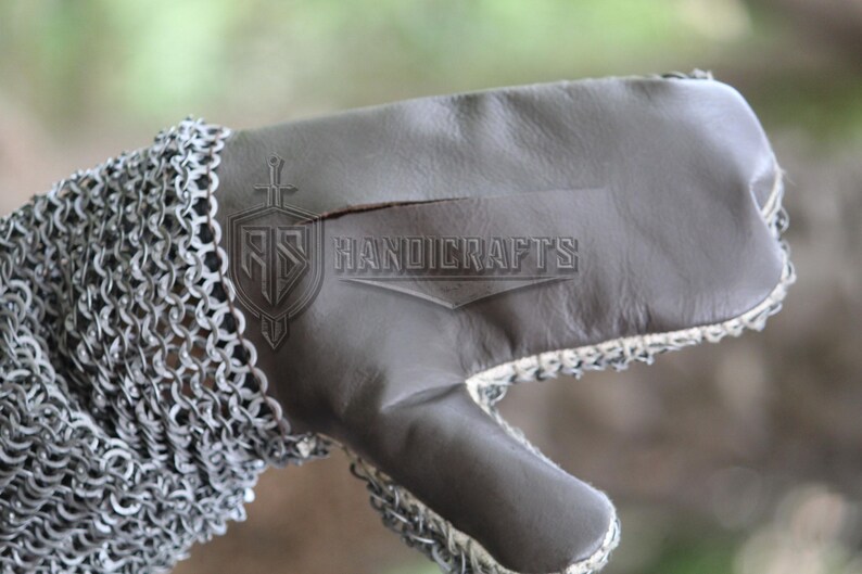 May include: Close-up of a medieval gauntlet. The gauntlet features a chainmail sleeve and a gray leather glove. The text "HANDICRAFTS" is visible on the leather. The gauntlet is designed for protection and historical reenactment.