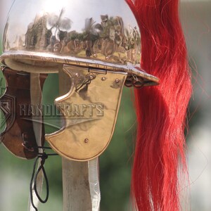 May include: A polished metal Roman helmet with a red plume. The helmet features a curved visor and cheek guards. The image also shows the text "HANDICRAFTS".