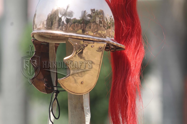 May include: A polished metal Roman helmet with a red plume. The helmet features a curved visor and cheek guards. The image also shows the text "HANDICRAFTS".
