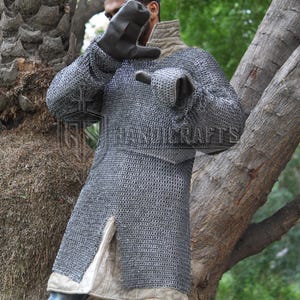 May include: A silver chainmail shirt with long sleeves and a split hem. The shirt is worn over a light-colored undergarment. The image shows a person wearing the chainmail shirt. The background includes trees and foliage.