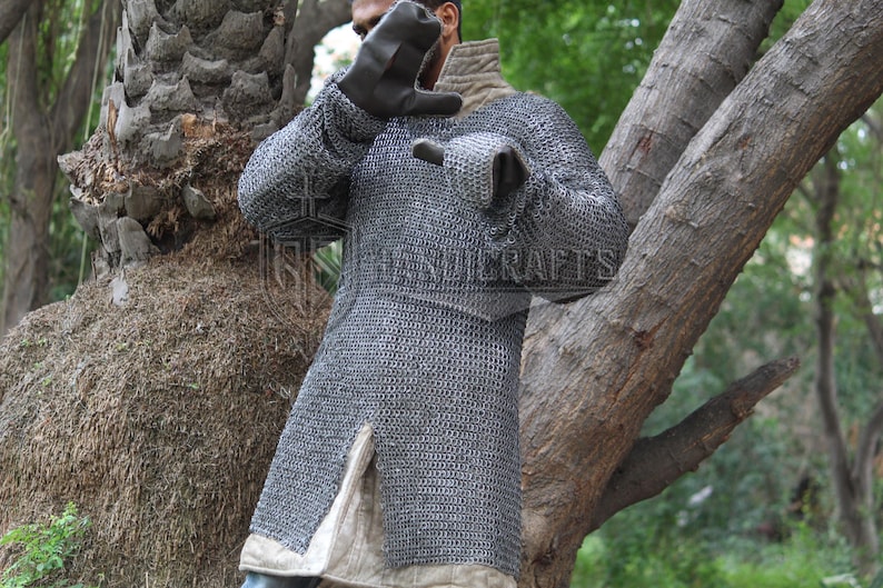 May include: A silver chainmail shirt with long sleeves and a split hem. The shirt is worn over a light-colored undergarment. The image shows a person wearing the chainmail shirt. The background includes trees and foliage.