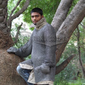 May include: A silver chainmail shirt with long sleeves and attached gloves. The shirt has a high collar and a light-colored trim. The person is wearing blue jeans. The background is a natural outdoor setting with trees.