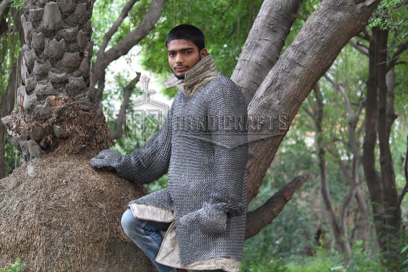 May include: A silver chainmail shirt with long sleeves and attached gloves. The shirt has a high collar and a light-colored trim. The person is wearing blue jeans. The background is a natural outdoor setting with trees.