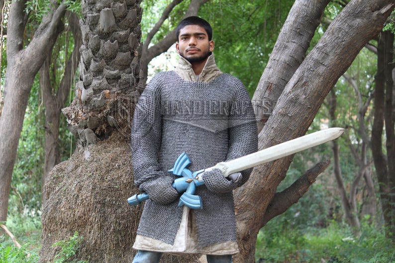 May include: A person wearing a chainmail shirt and holding a wooden sword with a blue hilt. The person is standing in a forest setting. The chainmail shirt is gray and the sword is white. The image has the text "HANDICRAFTS".