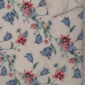 May include: White floral quilt featuring a repeating pattern of blue tulips, red and pink flowers, and green leaves. The quilt has a quilted design and a soft, plush texture. The overall theme is floral and botanical.