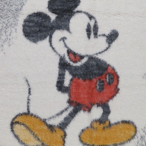 May include: A fuzzy, textured fabric featuring a graphic illustration of Mickey Mouse. The character is depicted in black, white, and red with large yellow shoes, set against a light, speckled background with a soft, plush, and tactile appearance.