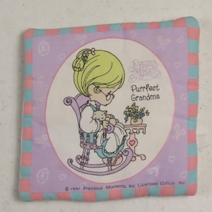 May include: Square, quilted Precious Moments pillow featuring a cartoon grandma in a rocking chair holding a cat. The pillow has a lavender center with the words "Purrfect Grandma." The border is pink and blue checkered. © 1997 Precious Moments, Inc.