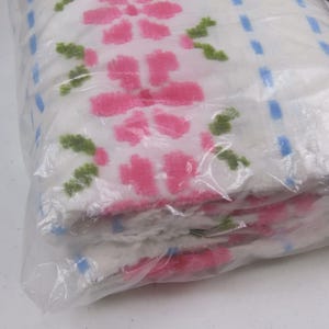 May include: A stack of white towels with a pink floral pattern and green leaves, visible through a clear plastic bag. Blue square accents are also present. The towels appear soft and fluffy.