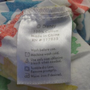 May include: A white fabric label featuring the Disney logo and care instructions. The label states "100% Cotton, Made in China, RN #117835." Washing instructions include machine wash cold, non-chlorine bleach, tumble dry low, and warm iron.