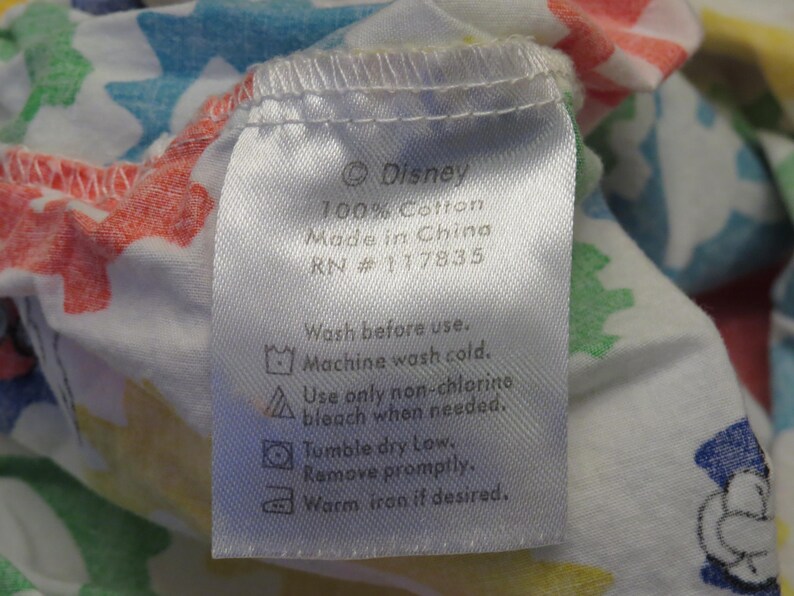 May include: A white fabric label featuring the Disney logo and care instructions. The label states "100% Cotton, Made in China, RN #117835." Washing instructions include machine wash cold, non-chlorine bleach, tumble dry low, and warm iron.