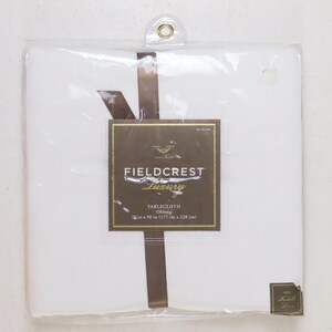 May include: A white Fieldcrest Luxury oblong tablecloth packaged in clear plastic with a brown ribbon. The label displays the text Fieldcrest Luxury, Tablecloth, Oblong, and 70 in x 90 in (177 cm x 228 cm). A small corner label indicates 100% Washable Linen.