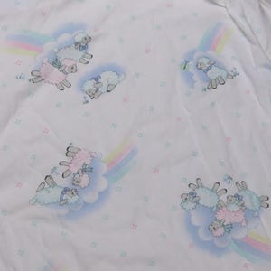 May include: White fabric with a repeating pattern of cartoon sheep, rainbows, and clouds. The sheep are in shades of pink, blue, and white, with pastel rainbows. Small, colorful square accents are scattered across the background.
