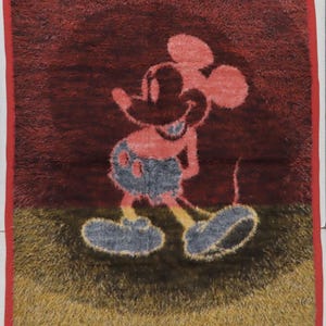 May include: A rectangular rug with a shaggy texture featuring a central graphic of Mickey Mouse. The background is divided into a dark red upper section and a golden-brown lower section. The rug is finished with a solid red fabric border trim.