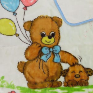 May include: A soft, white baby blanket featuring a brown teddy bear holding three balloons in blue, pink, and yellow. A smaller brown dog is next to the bear. The blanket has a blue border and a floral design at the bottom.