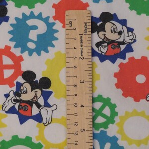 May include: White fabric with a repeating pattern of colourful gears and Mickey Mouse illustrations. The gears are in green, blue, yellow, and red. A wooden ruler shows measurements in inches and centimetres.