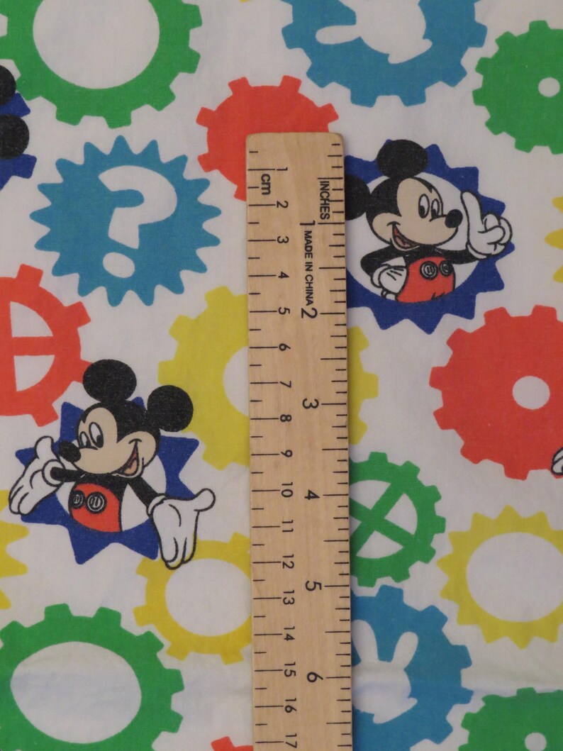 May include: White fabric with a repeating pattern of colourful gears and Mickey Mouse illustrations. The gears are in green, blue, yellow, and red. A wooden ruler shows measurements in inches and centimetres.
