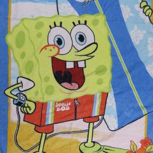 May include: A beach towel featuring a cartoon image of SpongeBob SquarePants in his swim trunks and flippers. The towel has a blue and yellow background with a surfboard that says "Bikini Bottom Boards".