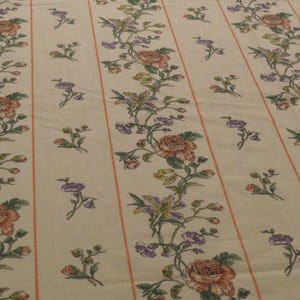 May include: Cream-coloured fabric with a repeating floral pattern. The design features vertical stripes with pink borders. Floral elements include roses, purple flowers, and green leaves. The overall aesthetic is vintage and floral.
