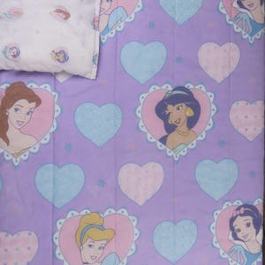 Peut inclure: Un ensemble de couette et d'oreiller lavande avec des motifs de cœurs et des illustrations de princesses Disney. La couette présente un motif répétitif de cœurs bleu clair et roses, chacun avec un portrait de princesse. L'oreiller présente des portraits de princesses.
