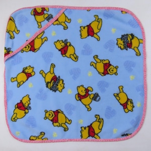 May include: A light blue baby blanket featuring a repeating pattern of the Winnie the Pooh character in various poses. The blanket has a red and white gingham trim and a hood. The blanket is made of a soft material.