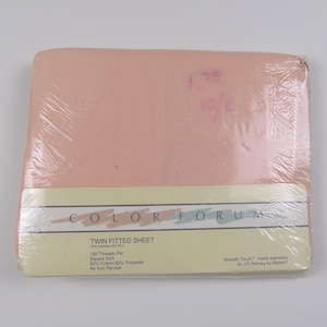 May include: A packaged twin fitted sheet in a light peach color. The sheet is wrapped in clear plastic. The label reads "COLOR FORUM" and "TWIN FITTED SHEET". The sheet is made of 50% cotton and 50% polyester.