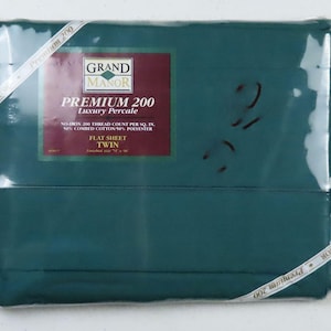 以下が含まれることがあります： 深いティール色のツインサイズのフラットシーツのパッケージ。パッケージには「GRAND MANOR PREMIUM 200 Luxury Percale」の文字と、糸数と素材構成に関する詳細が記載されたラベルが付いています。「Premium 200」と書かれた白いリボンがパッケージを包んでいます。