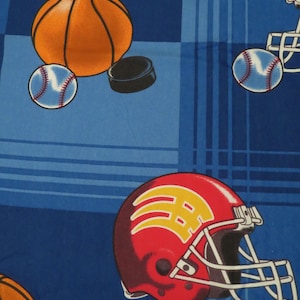 May include: Blue fabric with a pattern of sports equipment. The design includes an orange basketball, a baseball, a hockey puck, and a red football helmet with yellow accents. The background has a plaid design.
