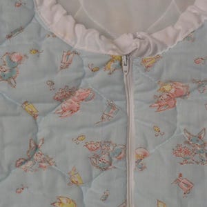 May include: A light blue quilted baby sleep sack with a white zipper and trim. The sleep sack is patterned with cartoon bunnies, gifts, and butterflies in pastel colors. The quilted design is visible throughout the fabric.