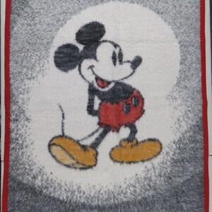May include: A rectangular fuzzy blanket featuring a central graphic of Mickey Mouse in black, white, red, and yellow. The character is set against a white circular background, surrounded by a grey textured border and a solid red fabric trim edge.