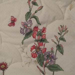 May include: A close-up of a white quilted fabric with a floral pattern. The design features red, purple, and green flowers, along with a small red butterfly. The background has a subtle polka dot pattern.