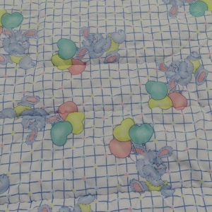 May include: A white baby blanket with a blue grid pattern and repeating illustrations of blue bunnies holding heart-shaped balloons in pastel colors. The blanket is quilted.