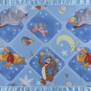 May include: A blue blanket featuring a pattern of Winnie the Pooh characters, including Pooh, Tigger, Eeyore, and Piglet. The blanket has a blue and white checkered border and is decorated with stars, honey pots, and bees.