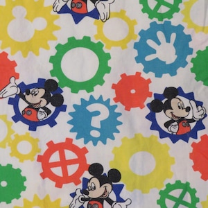 May include: White fabric with a repeating pattern of Mickey Mouse characters in blue starbursts, alongside colourful gear-shaped designs in red, yellow, green, and blue. The design is playful and vibrant.