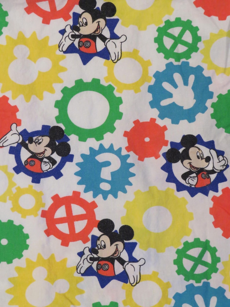 May include: White fabric with a repeating pattern of Mickey Mouse characters in blue starbursts, alongside colourful gear-shaped designs in red, yellow, green, and blue. The design is playful and vibrant.