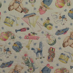 May include: A white fabric with a repeating pattern of vintage children's toys. The design includes teddy bears, toy blocks, drums, airplanes, and other classic playthings in soft pastel colors. Small blue snowflake accents are scattered throughout.