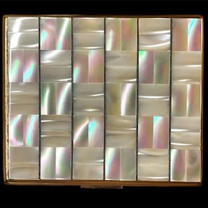 Può includere: Un astuccio compatto quadrato caratterizzato da una griglia di tessere in madreperla iridescente disposte secondo un motivo geometrico. La superficie presenta una finitura bianca e madreperlacea scintillante con sottili riflessi arcobaleno, incorniciata da un sottile bordo in metallo dorato.