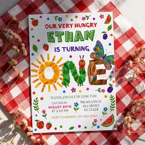 以下が含まれることがあります： イーサンの1歳の誕生日のカラフルな招待状。「Our Very Hungry Ethan is Turning One」のテキスト入り。太陽、イモムシ、蝶、果物のイラストがデザインされています。招待状は赤と白のチェック柄の背景にあります。