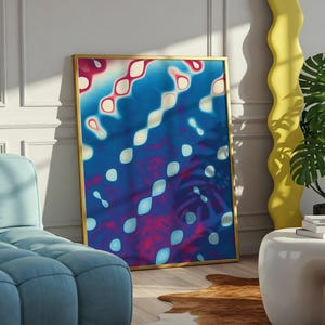 May include: Abstract art print in a gold frame. The artwork features a vibrant mix of blue, red, and white shapes. The print leans against a white wall, with a blue sofa and a yellow decorative element in the background.