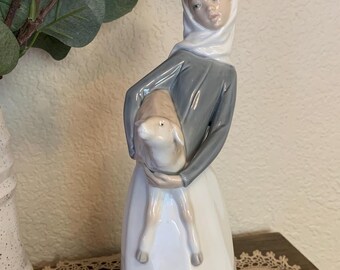 May include: A porcelain figurine of a young person in a white headscarf and gray dress, holding a lamb. The figurine has a detailed face and is set against a neutral background. The base of the figurine is a light gray color.