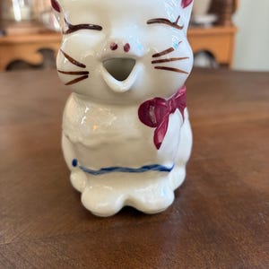 May include: A white ceramic creamer shaped like a cat with a smiling face, closed eyes, and brown painted whiskers. The cat features a dark red bow on its side and a thin blue horizontal line across its lower body, with an open mouth spout.