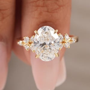 May include: A gold ring featuring a large oval-cut center stone held by prongs, flanked by smaller round-cut stones arranged in a cluster on each side of the band. The metal band is thin and polished, showcasing a classic prong setting design.