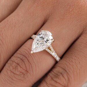 May include: A gold ring featuring a large pear-shaped center stone and a split shank band. The band is adorned with small, round, clear stones that extend along the sides, creating a sparkling effect against the polished yellow gold metal setting.