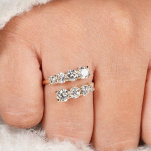 May include: A gold-toned ring with two bands, each adorned with multiple clear, round gemstones. The ring wraps around the finger, creating a unique and elegant design. The gemstones sparkle against the gold setting, creating a luxurious look.