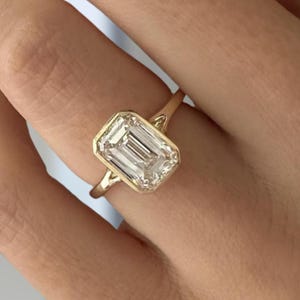 Op de afbeelding: Een gouden ring met een enkele, smaragdgeslepen heldere edelsteen in een gladde zetting. De ringband is dun en gepolijst, wat zorgt voor een strak ontwerp dat de rechthoekige vorm van de centrale steen en de warme kleur van het metaal benadrukt.