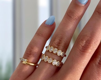 May include: Three gold rings featuring clear stones. Two rings are stacked on one finger, each set with a row of oval-shaped stones in a bezel setting. The third ring on the adjacent finger features a thin gold band with a small bar of clear stones.