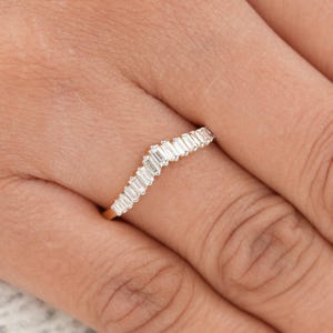 Puede incluir: Un anillo de oro curvado, engastado con una serie de piedras preciosas rectangulares y transparentes. El diseño del anillo presenta un patrón en forma de ola, creando un aspecto delicado y elegante. El anillo se muestra en un dedo.